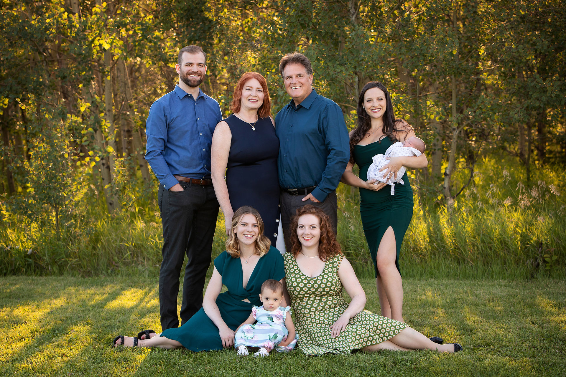 A family of seven, including two babies, poses outdoors on grass. Adults wear blue and green, surrounded by trees in sunlight. Smiling warmly.