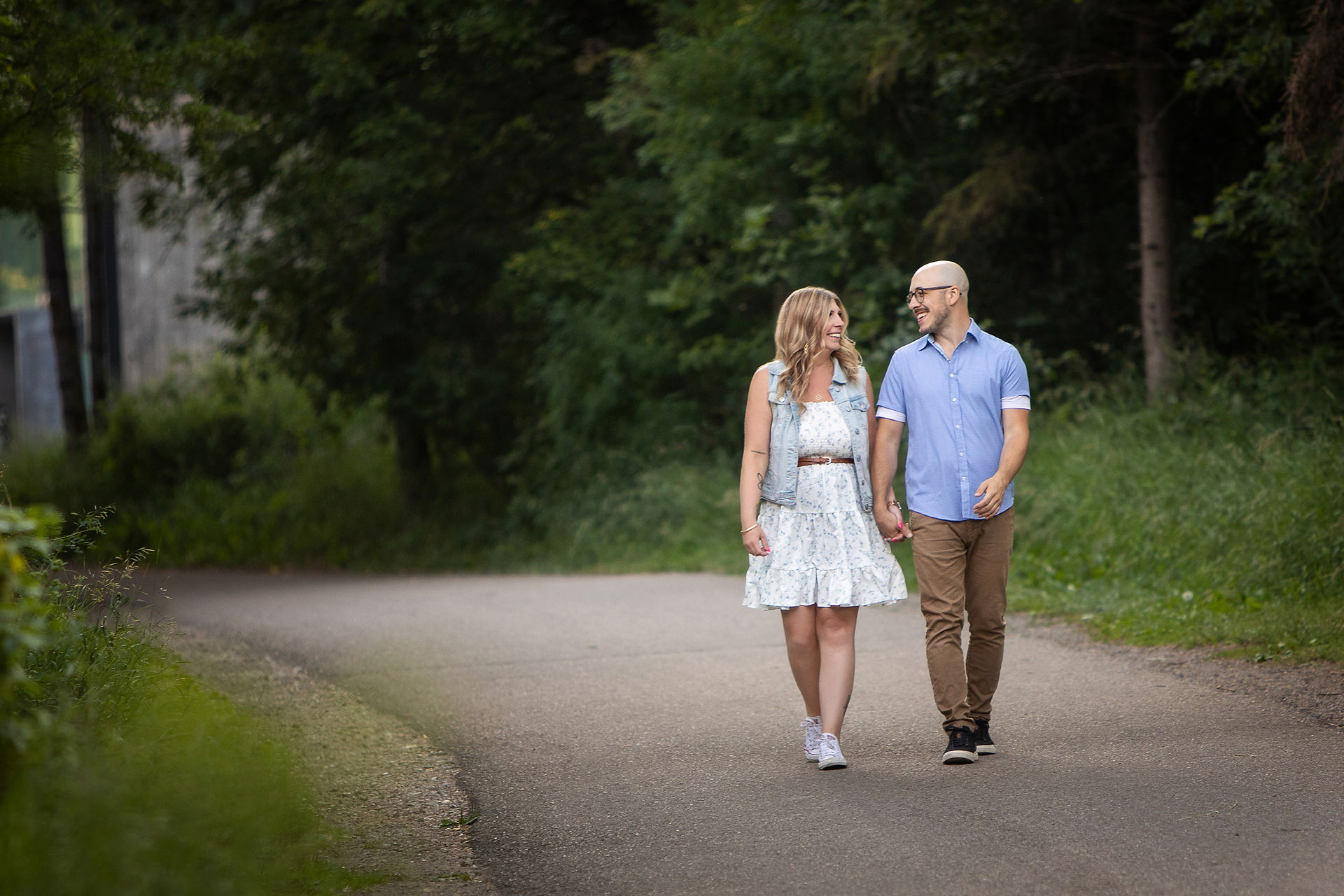 Couple walking hand in hand on a paved path in a lush green park, smiling at each other. Summer family photos