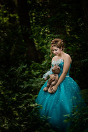 A young woman in a sparkly teal gown holds a teddy bear in a forest, exuding a serene, dreamy mood amidst lush green foliage. Themed grad photos Edmonton