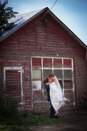 Bride in white dress lifted by groom in suit, smiling in front of rustic red barn. Joyful scene under a clear blue sky.