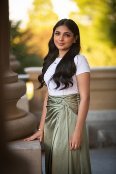 Woman in white top and green skirt smiles, standing by a column in a sunlit outdoor setting with blurred greenery background. Viewpoint Photography Edmonton