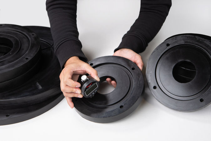Hands holding a black rubber disc with a gauge, inspecting its thickness. Additional black discs are nearby on a white surface.