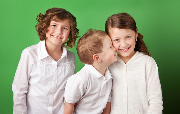 Three children in white shirts smiling against a green background. The boy leans in to whisper to the girl, creating a joyful mood. Children's Photography Edmonton