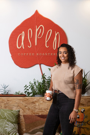 Smiling person holds coffee cup in front of a red sign reading Aspen Coffee Roasters. Plants and colourful cushions in the background.