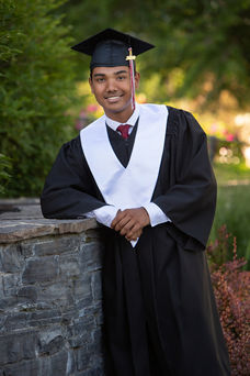 Graduate in cap and gown leans on stone wall, smiling. Black robe with white sash, red tie. Green, leafy background. Bright, celebratory mood.