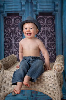 A smiling baby in jeans and a blue hat sits on a wicker chair with a decorative blue door backdrop,