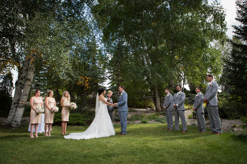 Bride and groom exchange vows outdoors, surrounded by bridesmaids in gold and groomsmen in gray suits. Lush greenery sets a serene mood. Viewpoint Photography Edmonton