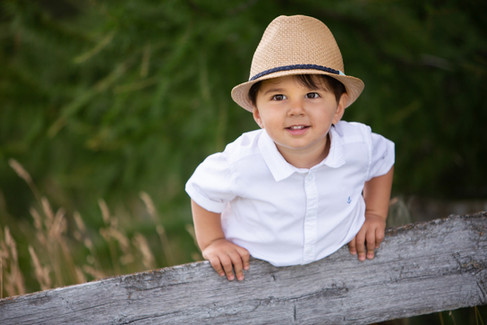 A child in a straw hat and white shirt leans on a weathered wooden fence, surrounded by green foliag