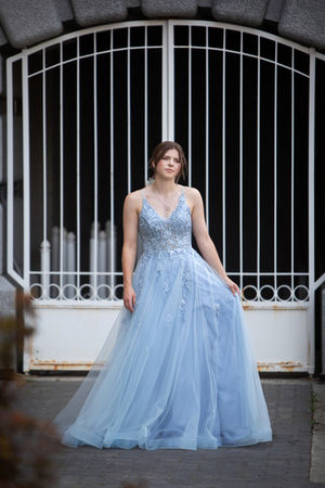 Young woman in a blue gown stands in front of a gated stone archway, holding grad dress fabric. The mood is serene, with intricate dress patterns. Grad photos Edmonton