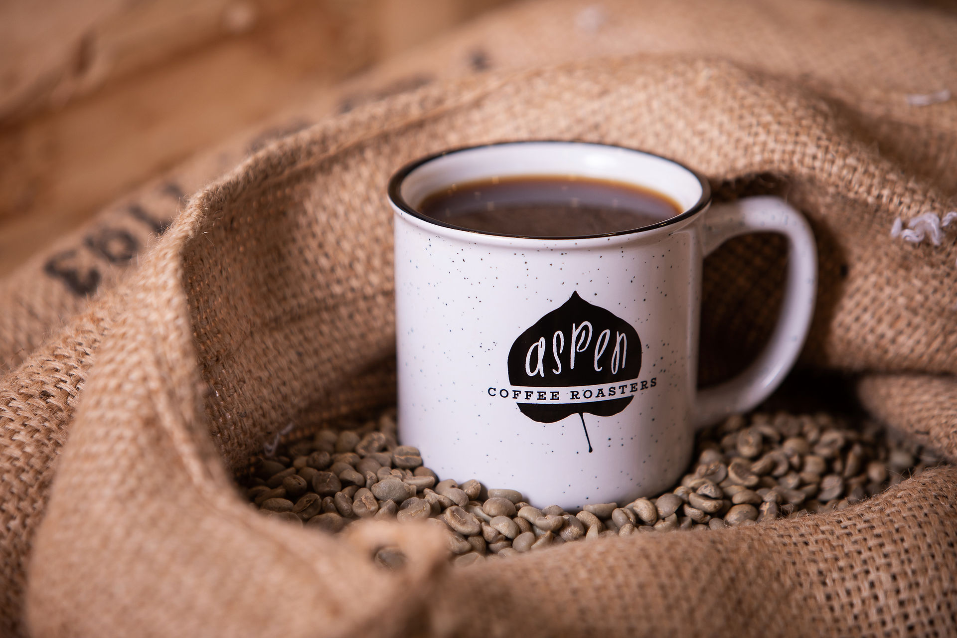 White mug with "Aspen Coffee Roasters" logo, filled with coffee, sits on green coffee beans atop burlap sacks, creating a cozy mood. - Edmonton Photographer