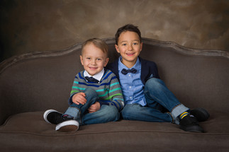 Two boys smiling, seated on a brown sofa. One wears a striped sweater, the other a bow tie and blazer. Warm, muted backdrop. Children's photography Edmonton