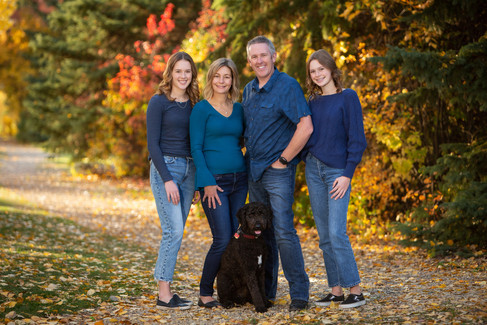Family of four and a black dog pose happily on a leaf-covered path, surrounded by colourful autumn trees, all wearing casual blue outfits. Family Photos Edmonton.