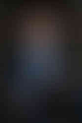 Man with curly red hair in a blue suit sits on wooden steps, smiling warmly. Wearing glasses, with green foliage in the background.