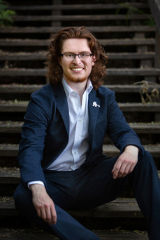 Man with curly red hair in a blue suit sits on wooden steps, smiling warmly. Wearing glasses, with green foliage in the background.