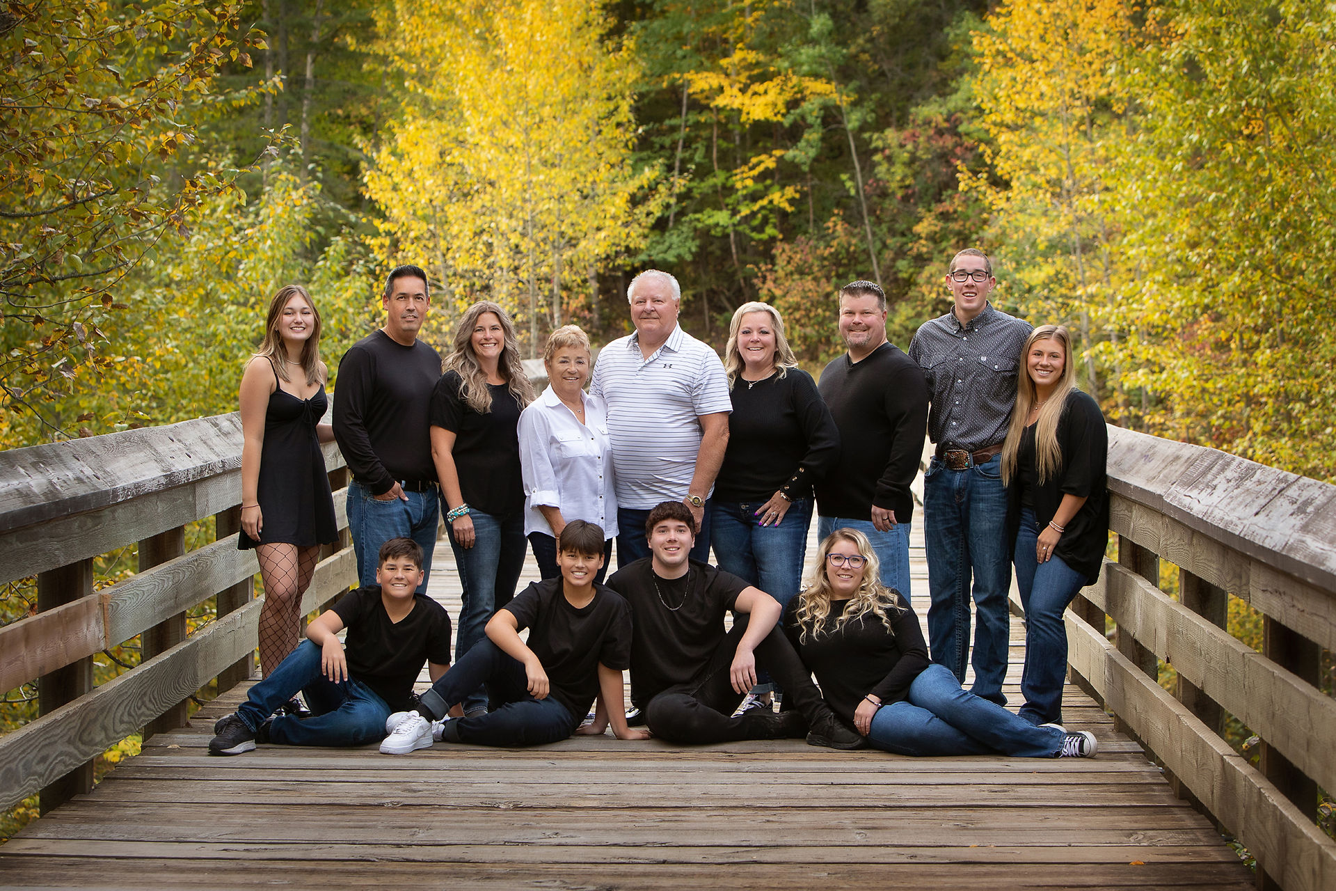 A group of 13 people on a wooden bridge, surrounded by autumn trees. They are smiling and wearing casual black and blue clothing.