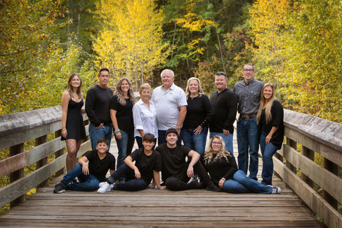 A group of 13 people on a wooden bridge, surrounded by autumn trees. They are smiling and wearing casual black and blue clothing.