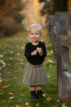 Young child smiling, holding a leaf, stands next to a wooden fence on a grassy path. Autumn leaves scattered, warm colours in background. Children's photography Edmonton