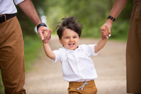 Child in white shirt and brown pants joyfully walks, holding hands with two adults on a path surroun