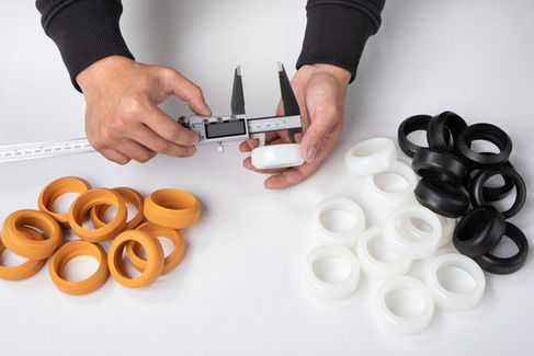Hands using calipers to measure a white ring. Orange, white, and black rings scattered on a white surface, creating a precise work mood.
