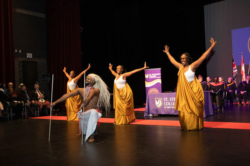 Dancers in golden outfits perform joyfully on stage with a kneeling figure in traditional attire. "St. Stephen's College" banner visible. Viewpoint Photography Edmonton