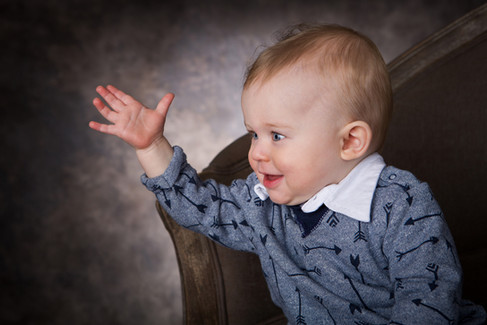 A smiling baby in a blue patterned sweater raises their hand, sitting on a brown chair against a blu