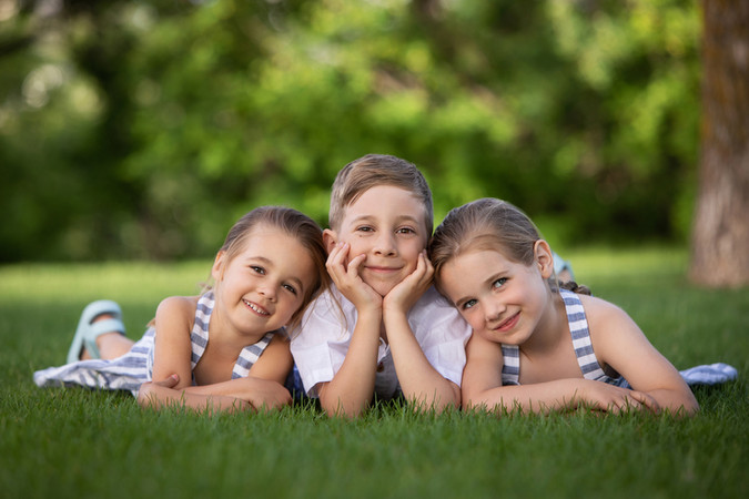 Three smiling children lie on grass, heads resting on hands, wearing striped outfits. Background is a blurred green park setting. Photography Edmonton