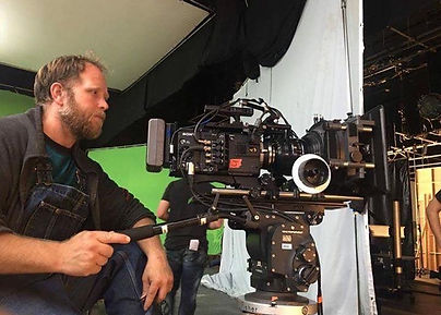 In the image, Robert is seated behind a large, professional RED camera on the set of a studio music video. He is focused on the shot, adjusting the camera's settings while sitting near it. Behind him, a large green screen covers the background, indicating that special effects or digital backgrounds will be added later. The setting is a professional studio environment, with lighting equipment and other production gear surrounding him, capturing the essence of a film director at work.