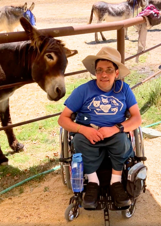 Man in wheelchair wearing a blue animal shirt and beige hat, smiling beside a donkey at Rancho Burro Donkey Sanctuary. Outdoor setting with more donkeys in the background.