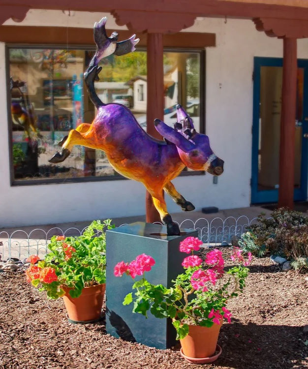 Colorful sculpture of a purple and orange animal with a bird on its tail, surrounded by potted pink flowers in a garden setting by artist Barbara Meilke Fine Art.