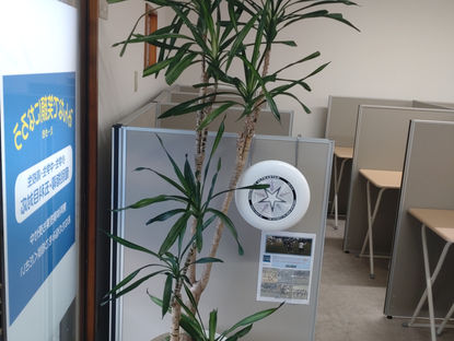 教室の仕切り席の間に大きな観葉植物のドラセナが立ち、左に教室の入り口、応援先のアルティメットグループの告知が見える静かな室内。