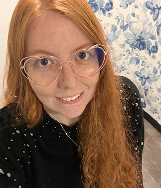 Red-haired woman with heart-shaped glasses smiling against blue floral background.