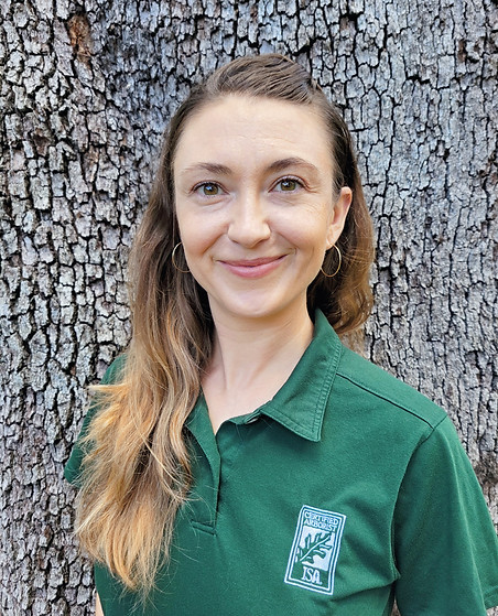 A profile picture of a caucasian woman with long brown hair and a Certified Arborist green polo on. In the background is tree bark