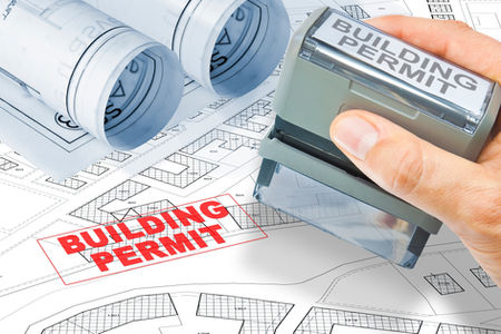 Hand stamping "BUILDING PERMIT" in red on architectural blueprints. Rolled plans and grid patterns in background, conveying approval.