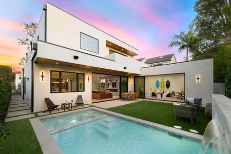 A luxurious modern home in North Vancouver with a sleek white exterior, large glass windows, and an open-concept indoor-outdoor living space. The backyard features a stylish swimming pool, manicured lawn, and cozy outdoor seating areas. The warm sunset sky adds to the elegant and inviting atmosphere.