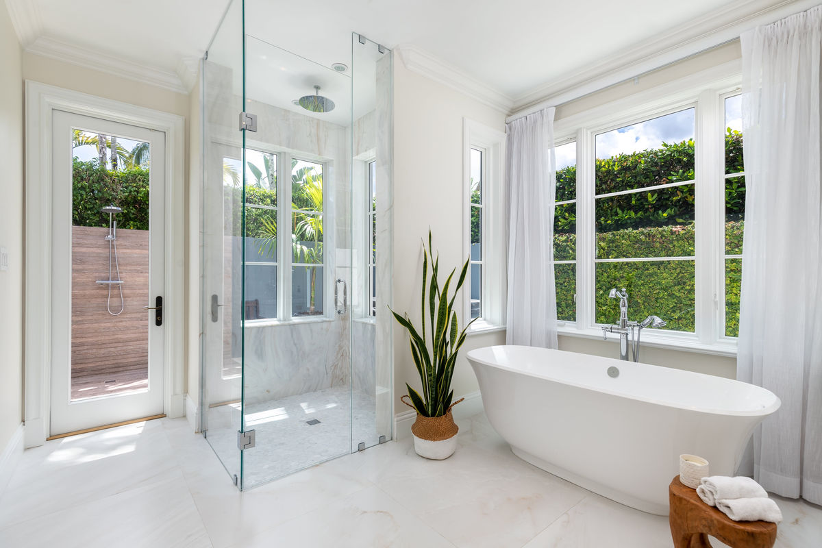 Luxurious bathroom with white bathtub, glass shower, large windows, and potted plant. Bright, airy ambiance with green garden view.