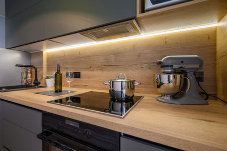 A modern kitchen with a sleek wood countertop and backsplash, featuring an induction cooktop with a stainless steel pot, a built-in oven, under-cabinet LED lighting, and stylish dark cabinetry. A stand mixer, wine bottle, and glass are neatly placed on the counter, enhancing the warm and contemporary design.