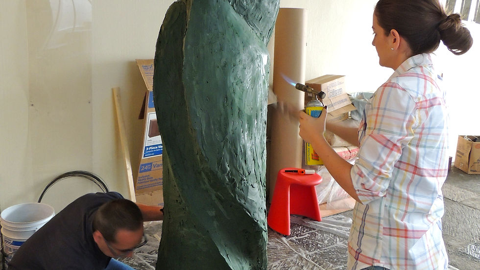 Two people work on a tall green sculpture in a workshop; one sprays it with a torch while the other kneels to clean the base.