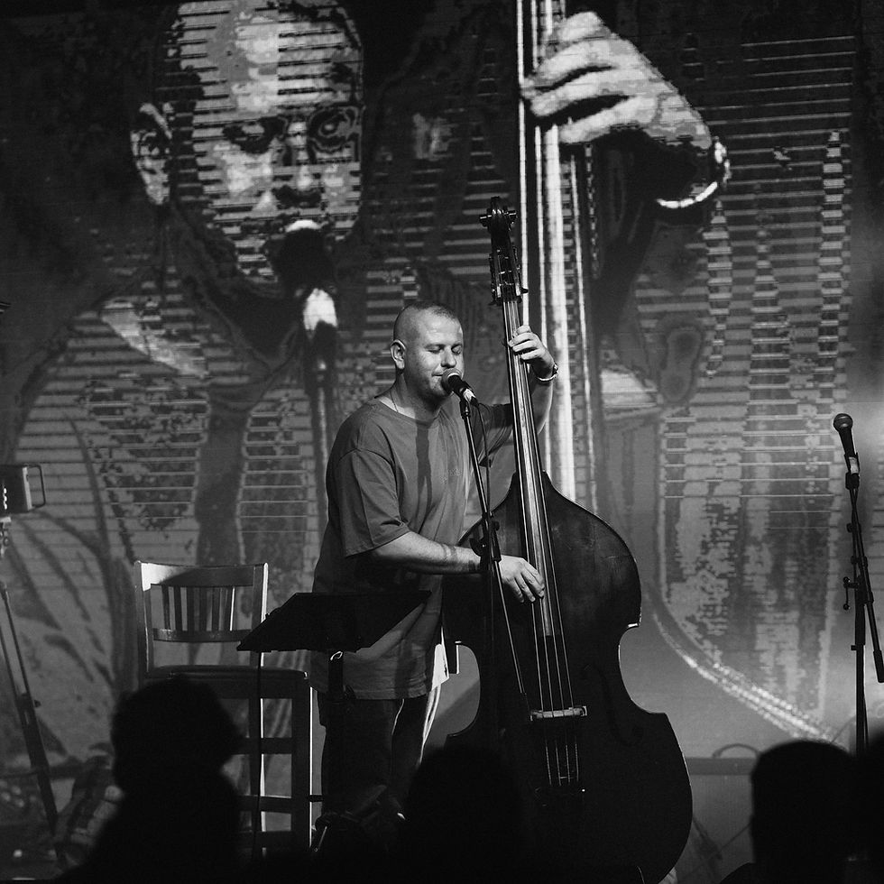 Black-and-white stage performance of a man singing into a microphone while playing upright bass, with a giant face backdrop.