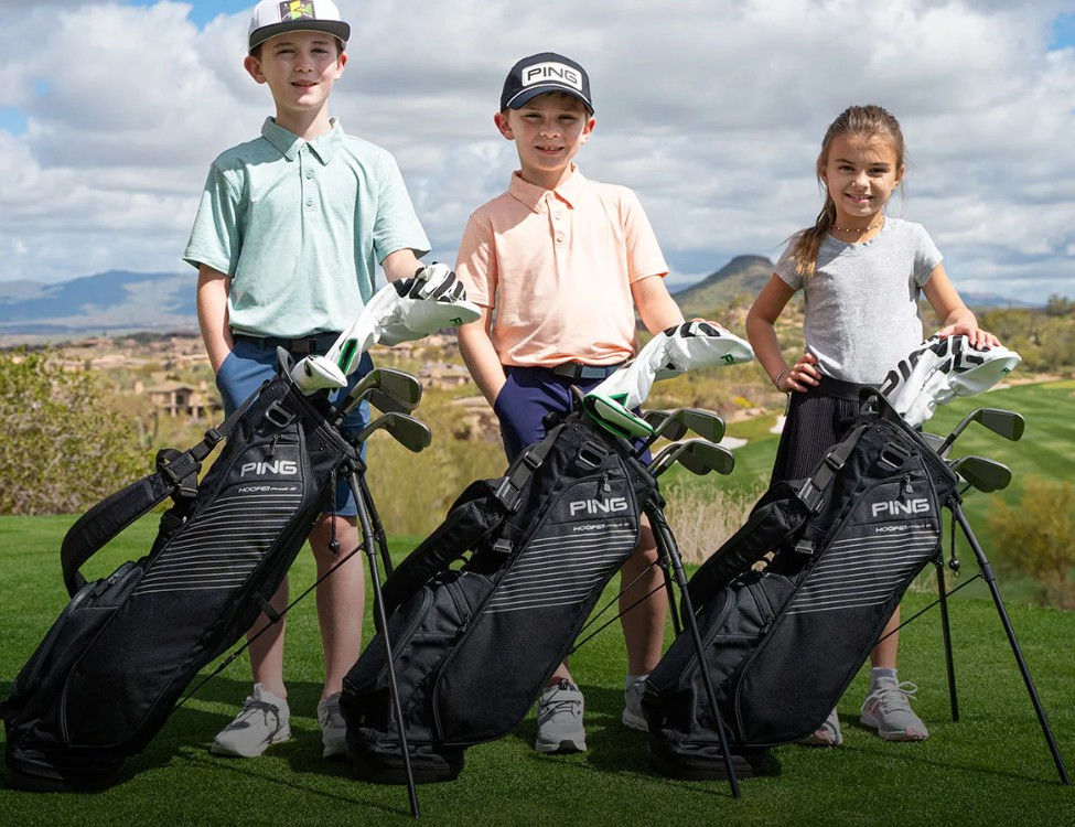 Three junior golfers with Ping Prodi G stand bags on a sunlit fairway, desert landscape behind them.