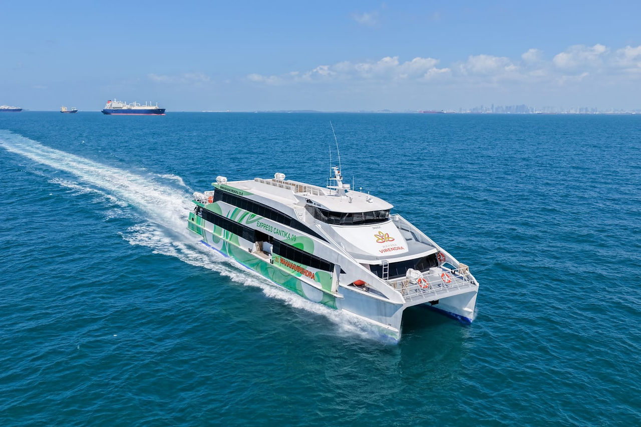 The Express Cantika fast boat catamaran operated by Wahana Virendra cutting through bright blue water at speed, with a large cargo vessel visible on the horizon
