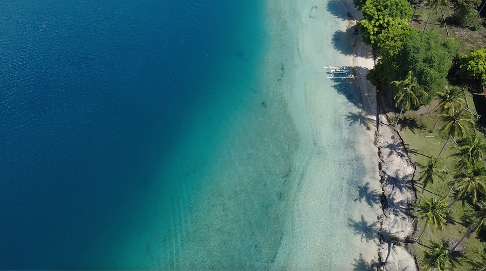 Aerial drone view of Gili Asahan’s untouched coastline with crystal clear turquoise water, white sand, palm trees, and a traditional boat resting near the shore