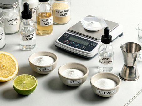 A white countertop displays three glass jars labeled "LACTIC ACID," "ACETIC ACID," and "ASCORBIC ACID," a digital scale with white powder, an amber dropper bottle labeled "ACID SOLUTION," a beaker, a metal jigger, a bar spoon, half a lemon, half a lime, and three ceramic bowls labeled "CITRIC," "MALIC," and "TARTARIC