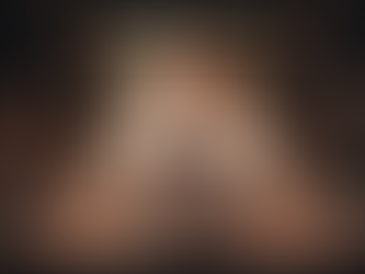 A professional male bartender with a beard, wearing a white shirt and a leather-strapped apron, stands behind a dark wooden bar counter. He is focused on a laptop screen placed in the center of the bar, surrounded by a large and organized variety of polished glassware, including wine glasses, coupes, and tumblers.