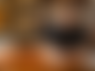 A smiling, bearded male bartender wearing a denim apron, leaning over a wooden bar counter and writing notes in a sketchbook. A classic Negroni cocktail with an orange peel garnish and professional bar tools are visible on the counter against a background of well-stocked liquor shelves.