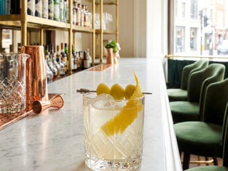 A clear, refreshing cocktail served in a faceted rocks glass on a white marble bar counter. The drink is garnished with three bright green olives on a silver cocktail pick and a long, elegant lemon twist. The background features a high-end, sunlit bar with polished brass shelving filled with bottles and forest-green velvet chairs near a large window.