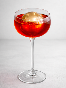 Bright red cocktail served in a clear coupe glass with a large gold spherical chiller resting on the surface, set against a clean light gray background.