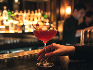 A close-up of a vibrant red Cosmopolitan cocktail served in a sleek coupe glass on a dark, reflective bar counter. A woman's hand with a delicate ring is reaching to pick up the drink. The background shows a dimly lit, sophisticated cocktail bar