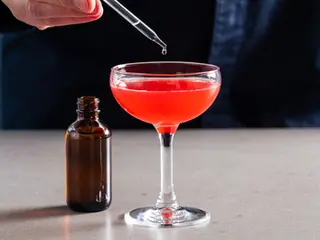 Close-up of a bartender's hand using a dropper to add a drop of 20% Saline Solution to a pink cocktail in a coupe glass. A labeled bottle of the saline solution sits on the counter next to the drink
