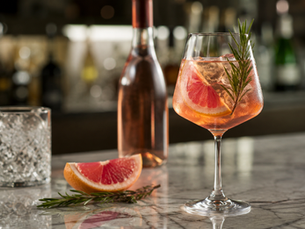 Rosé wine cocktail in a stemmed glass with grapefruit slice, rosemary garnish, ice, and condensation on a marble bar counter, with a rosé wine bottle and bar background softly blurred.