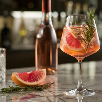 Rosé wine cocktail in a stemmed glass with grapefruit slice, rosemary garnish, ice, and condensation on a marble bar counter, with a rosé wine bottle and bar background softly blurred.